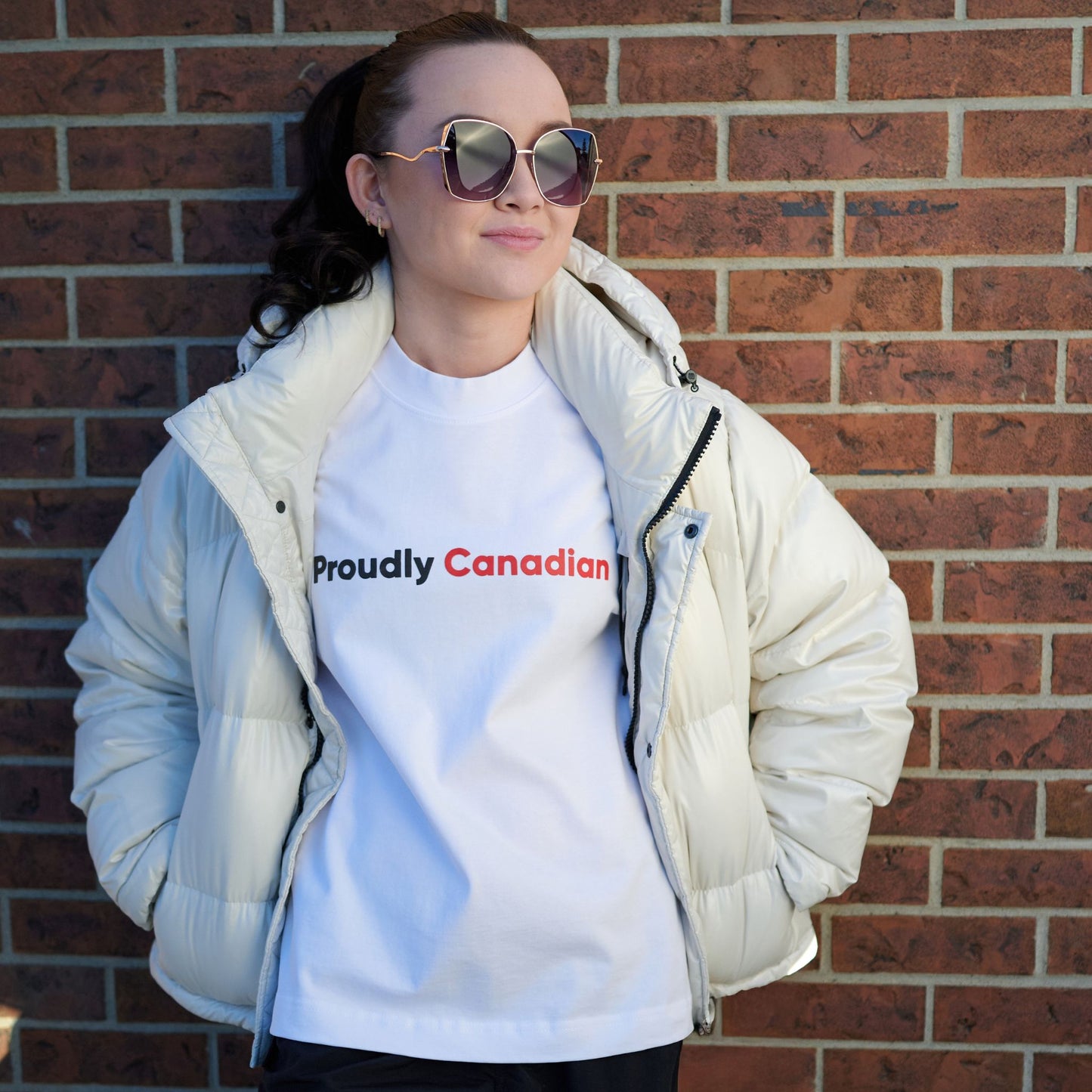 Female model wearing Proudly Canadian Women's T-Shirt in premium white cotton with raised black Proudly and red Canadian typography pairing with a winter coat in an outdoor urban setting
