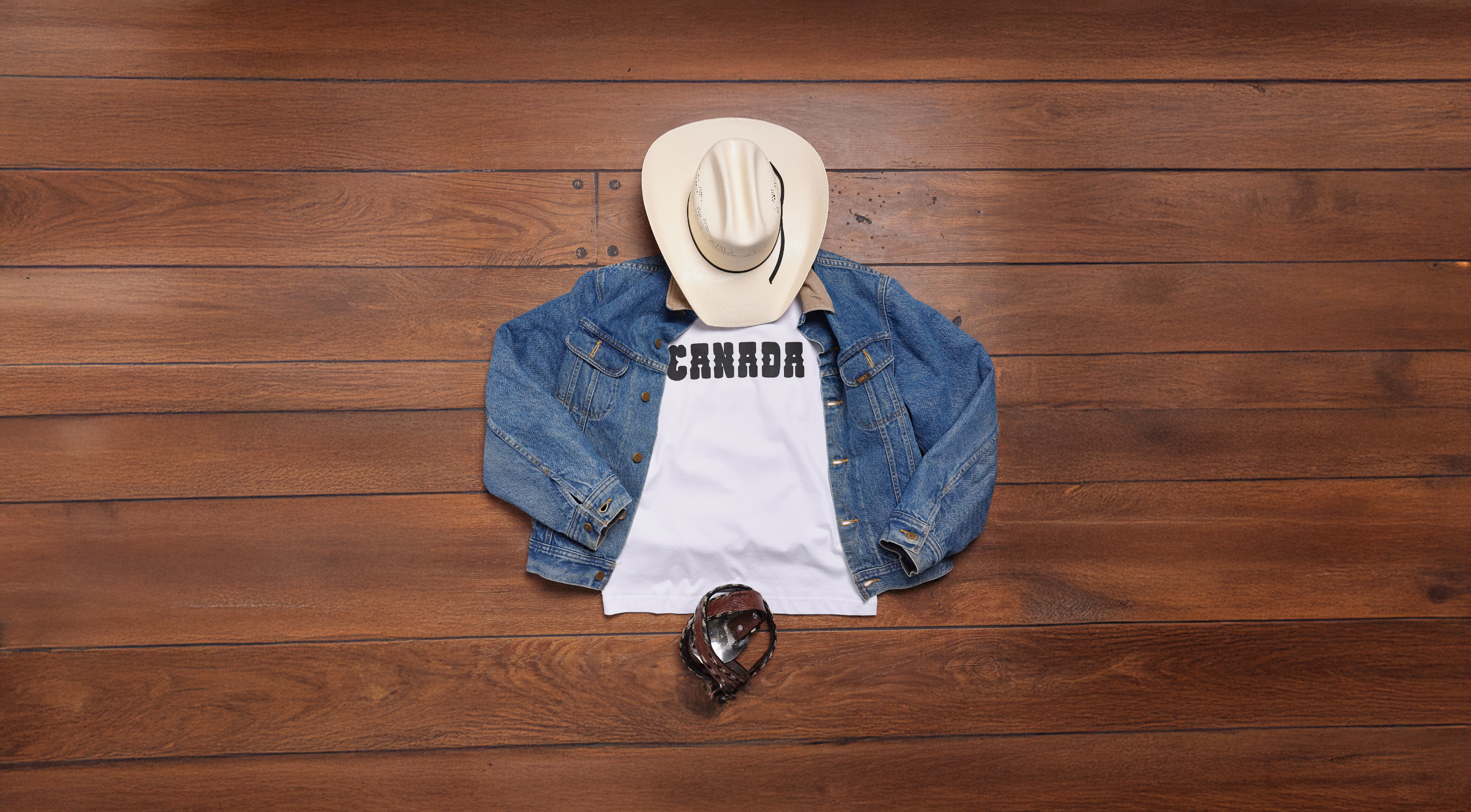 White Kermodee t-shirt with bold"CANADA" print styled with a denim jacket, cowboy hat, and belt on rustic wood - a nod to Canadian Western heritage.